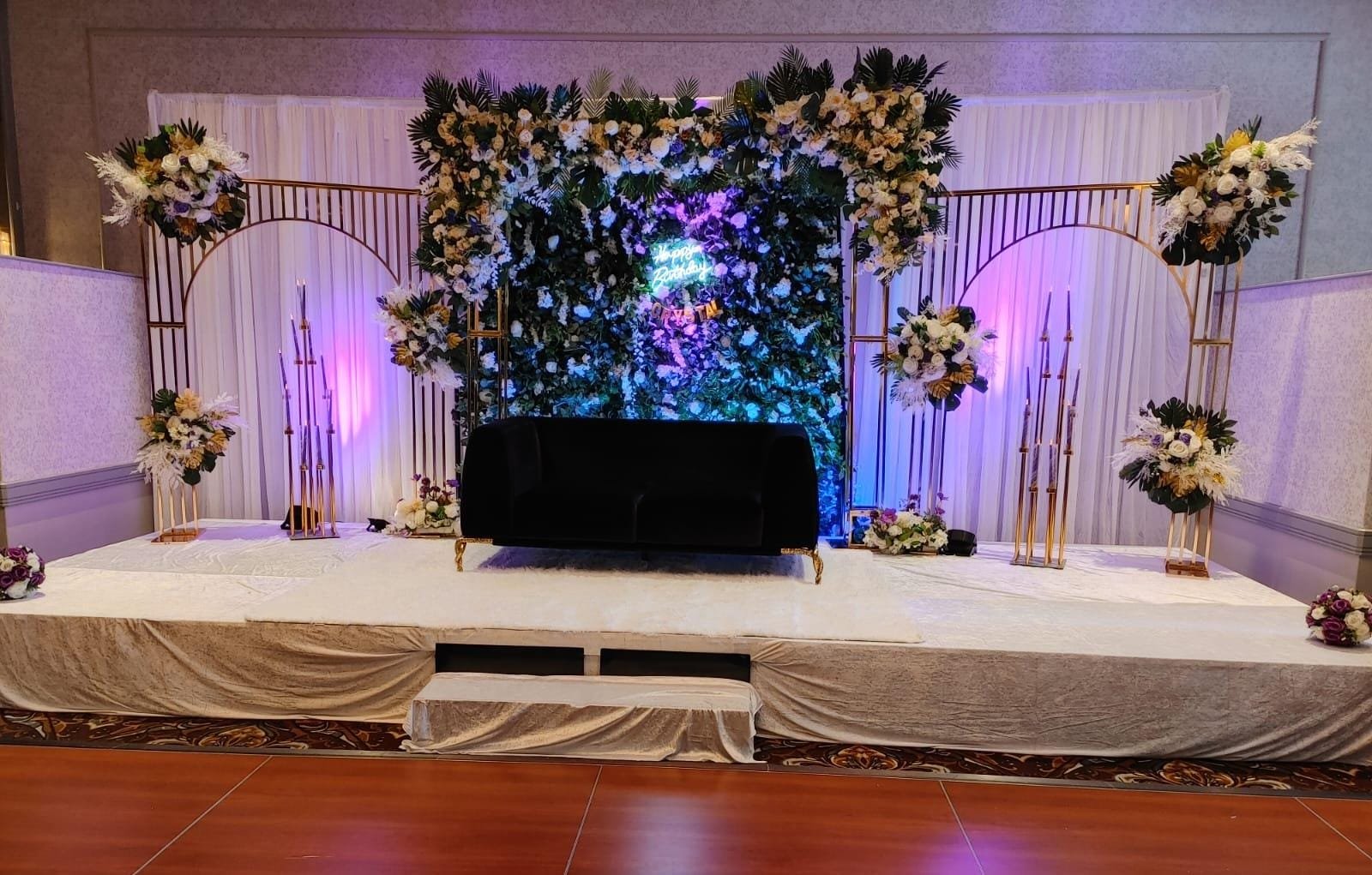 Birthday stage with floral wall, gold arches, candelabras and black velvet sofa